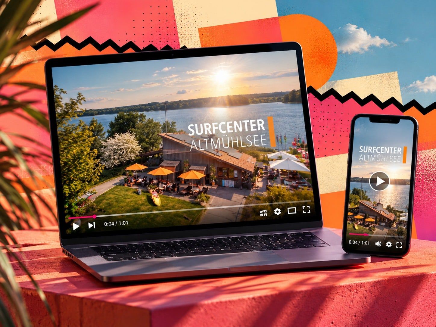 Laptop und Smartphone mit dem Imagefilm „Surfcenter Altmühlsee“: Luftaufnahme des Holzgebäudes mit orangen Sonnenschirmen am Seeufer bei Sonnenuntergang, vor buntem Hintergrund in Pink und Orange